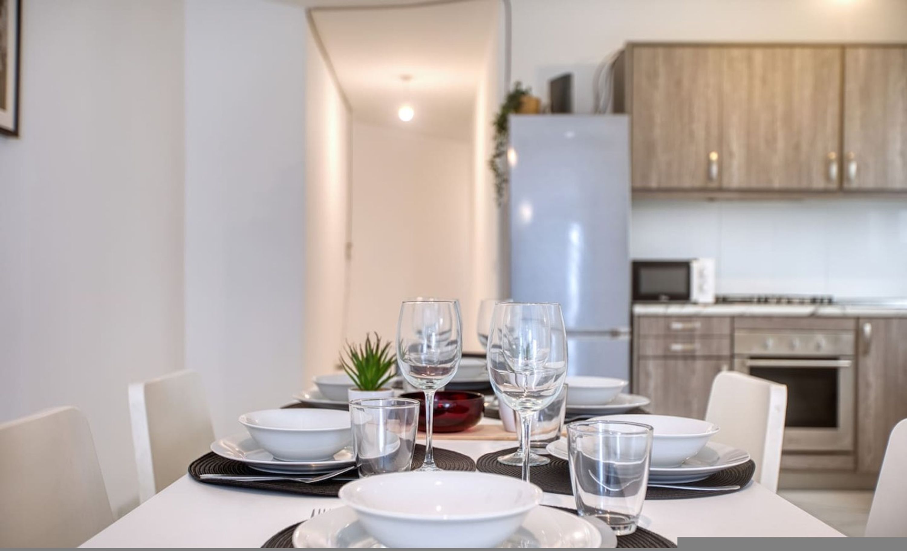 A dining table set with white plates, clear glasses, and a red bowl, with a kitchen in the background.