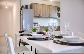 Un comedor con una mesa blanca dispuesta para cuatro personas, con platos, tazones, cubiertos y vasos. Hay plantas en macetas sobre la mesa, y se ve una cocina al fondo.