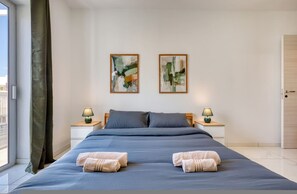Una habitación de hotel con una cama grande, dos mesitas de noche con lámparas, dos cuadros abstractos en la pared y vistas por la ventana.