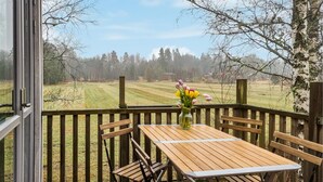 A wooden deck with a table and chairs, a vase with flowers, and a view of a grassy field and trees. - Nice home in Alingsås (Alingsås)