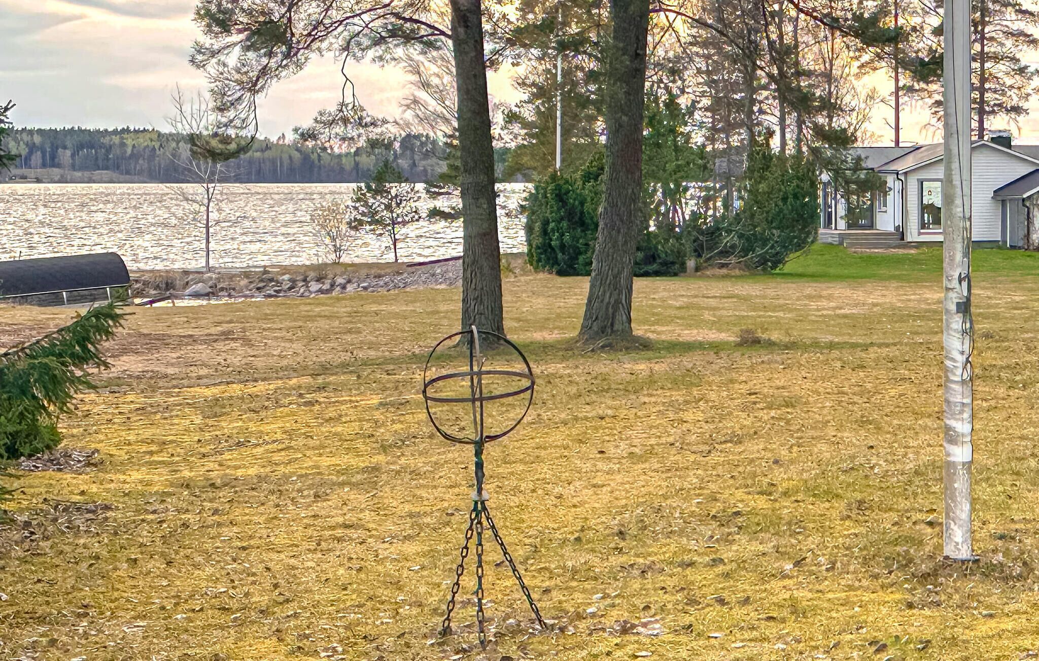 Una zona verda amb una escultura de metall, arbres i un cos d'aigua al fons.