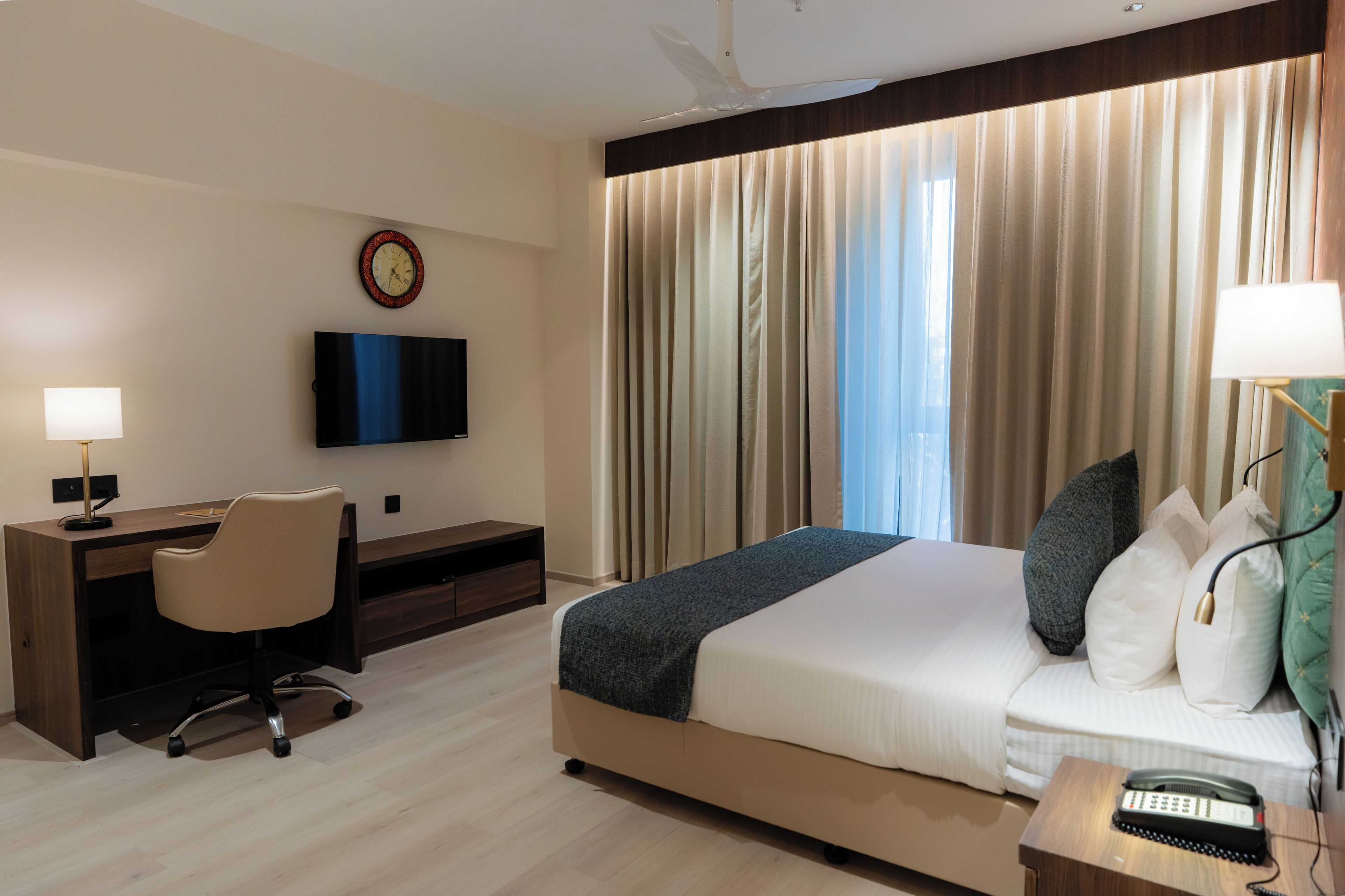 Una habitación de hotel con cama, escritorio, silla, televisor, cortinas y un reloj.