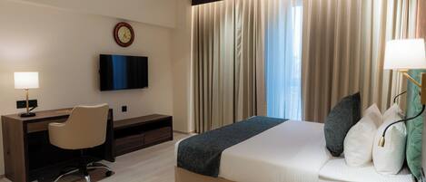 Una habitación de hotel con cama, escritorio, silla, televisor, cortinas y un reloj.