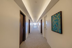 Um corredor comprido e estreito com portas em ambos os lados, chão coberto de carpete e teto com iluminação embutida.