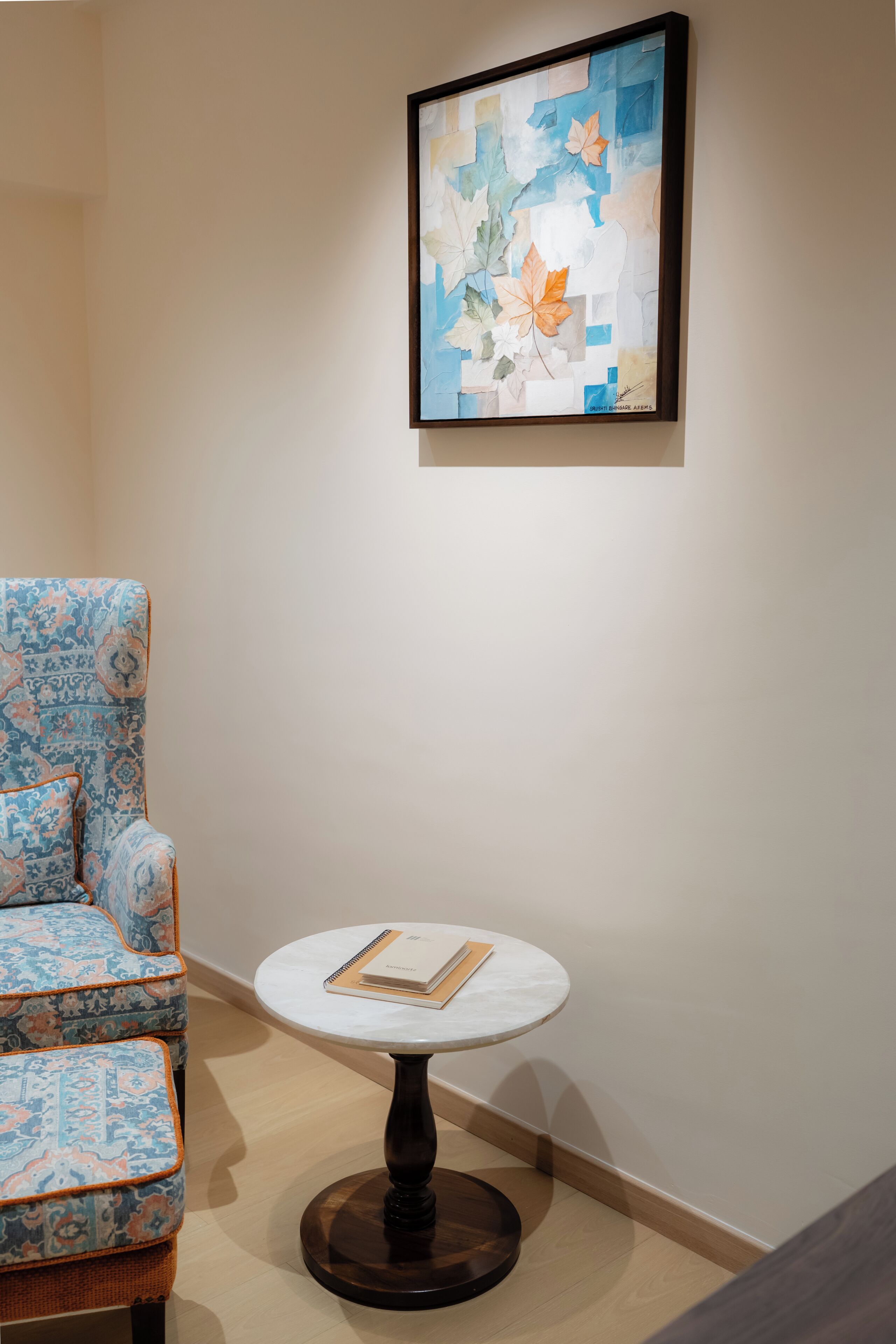 A room with a patterned armchair, a round table with a book, and a framed abstract painting on the wall.