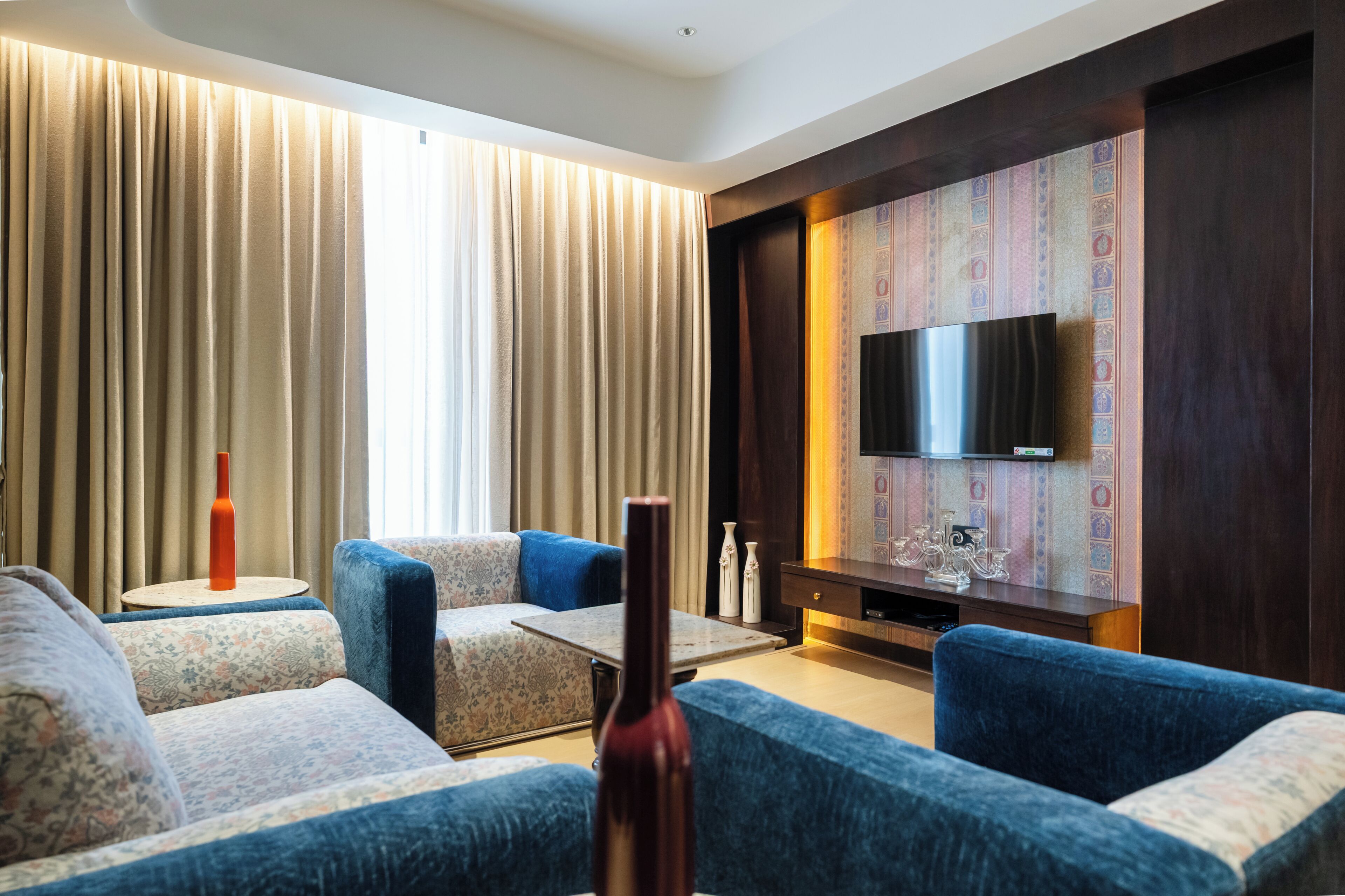 A modern living room with a sofa set, a coffee table, a flat-screen TV mounted on a patterned wall, and a large window with curtains.