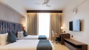 A hotel room with two beds, a desk, a chair, a TV, and a ceiling fan. - Akaya Elegance (Ahmednagar)