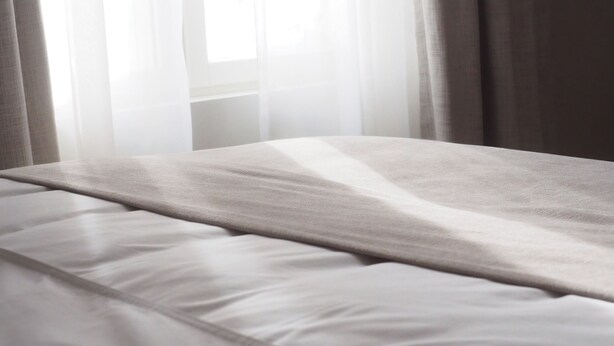 A bed with a white sheet and a blanket, curtains partially drawn, and a window in the background. - Sugar Factory Apartments (Kotka)