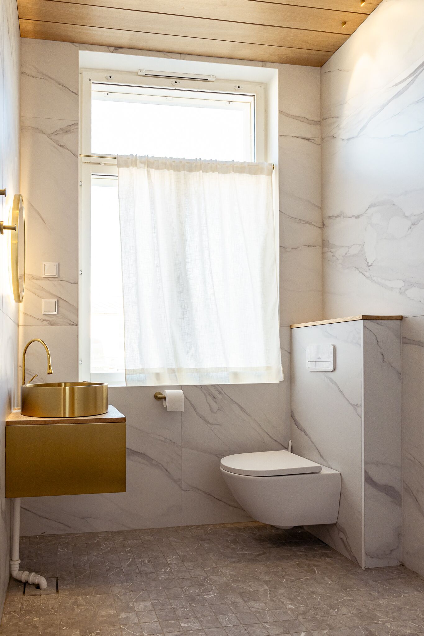 Een badkamer met een marmeren wand, een gouden wastafel, een raam met gordijn en een toilet.