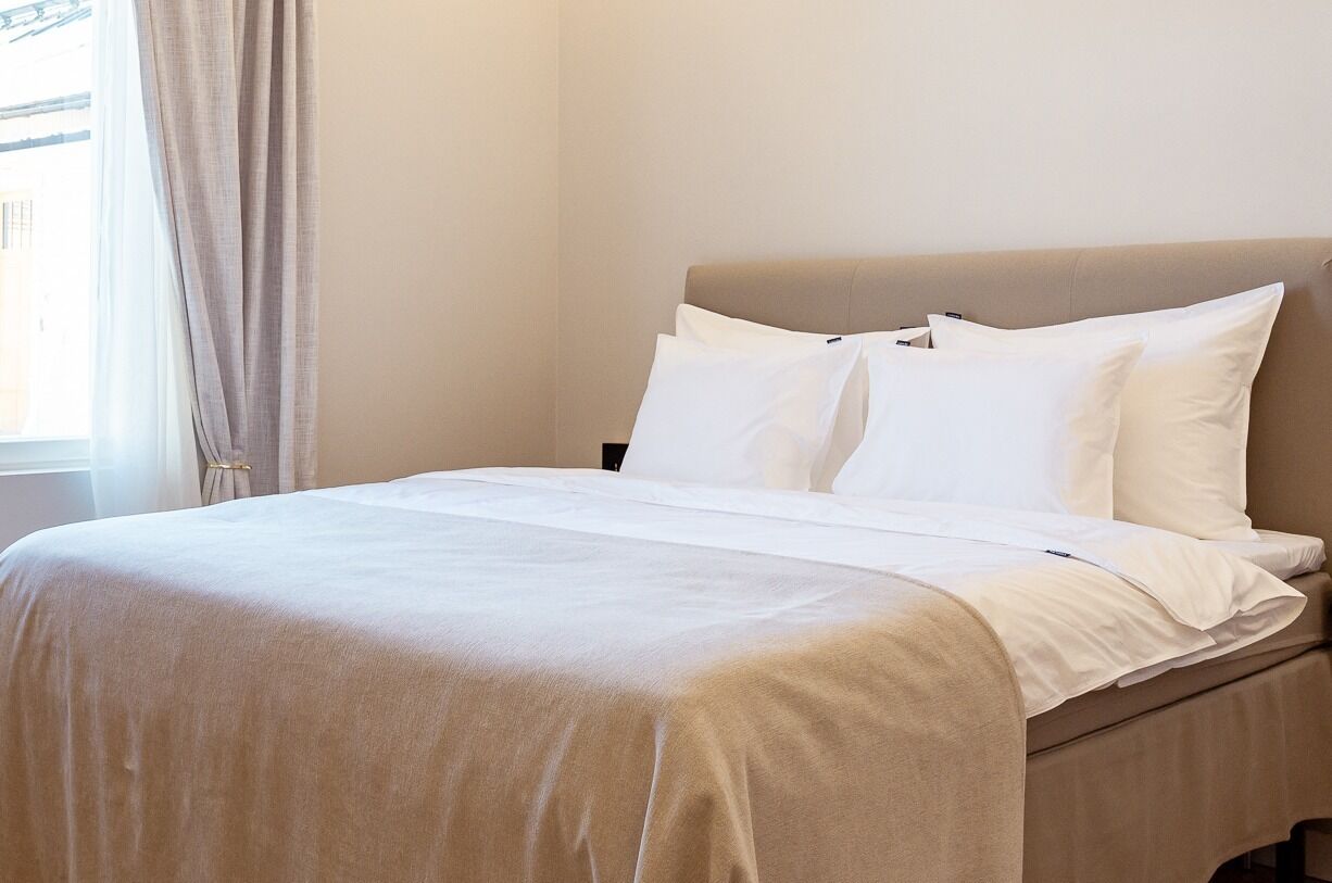 Una cama con sábanas blancas, cabecera color beige y una ventana con cortinas.