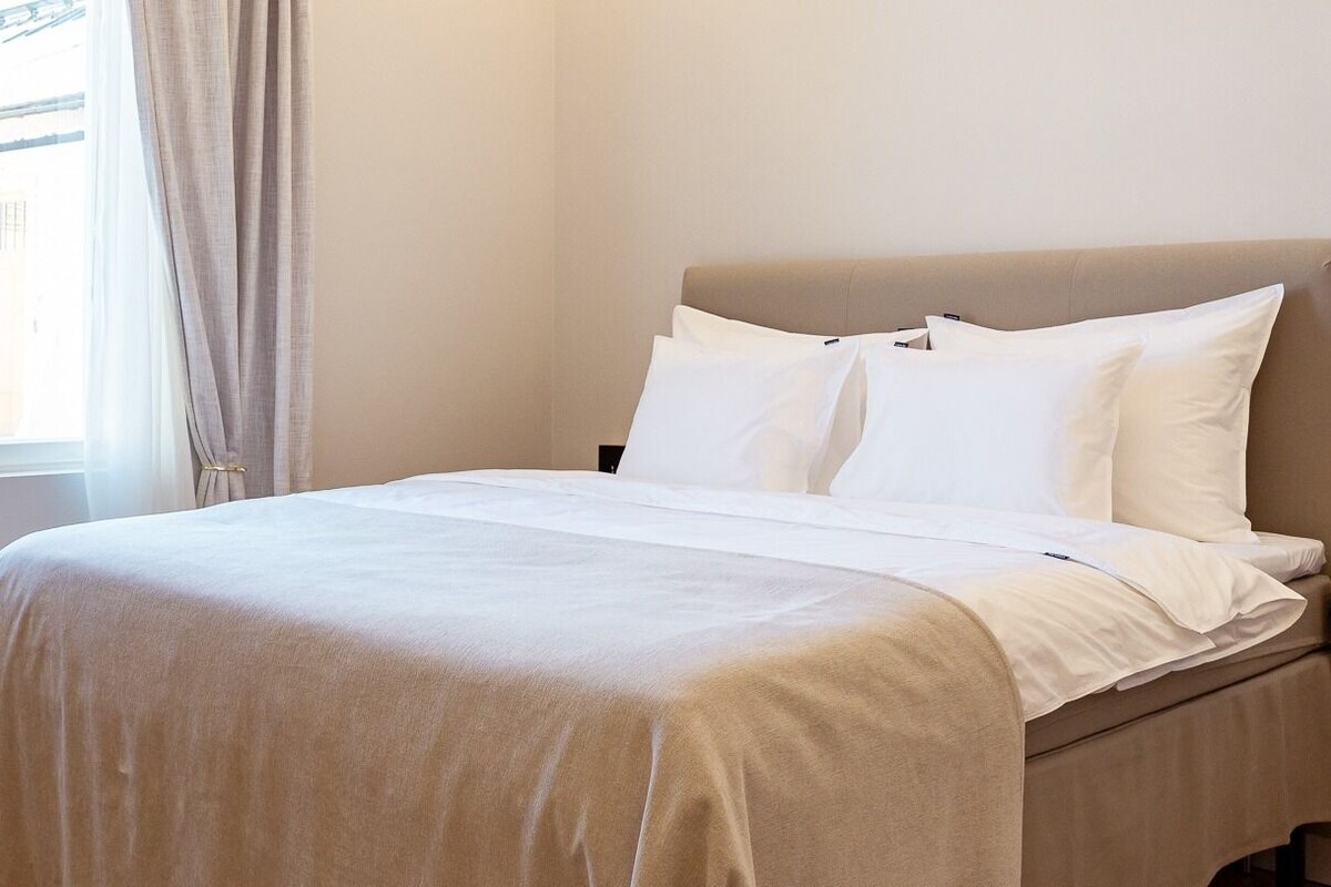 Ein Bett mit weißen Bettwäsche, einem beige-farbenen Kopfteil und einem Fenster mit Vorhängen.