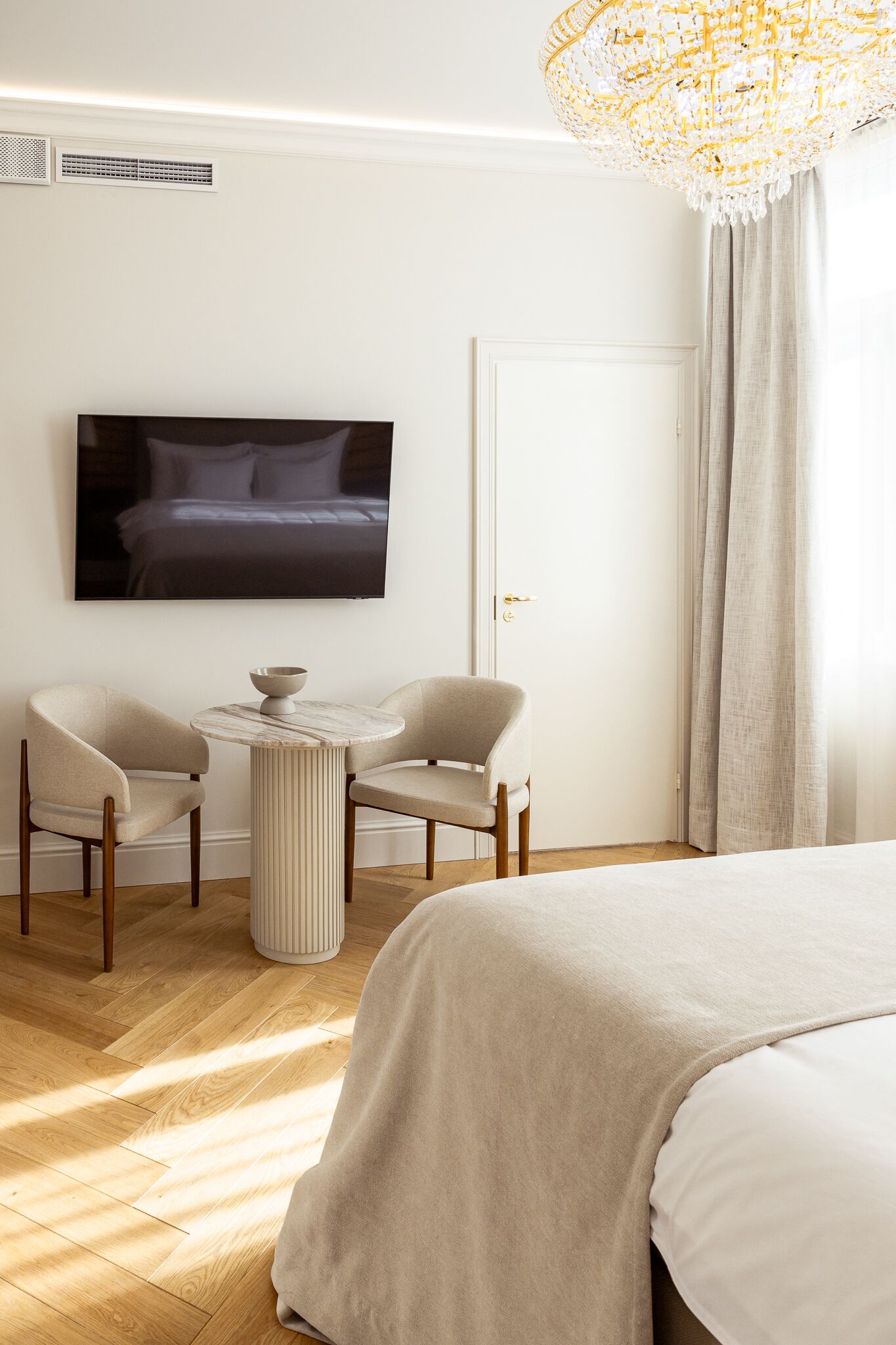 A hotel room with a bed, two armchairs, a small round table, a television mounted on the wall, and a large chandelier.