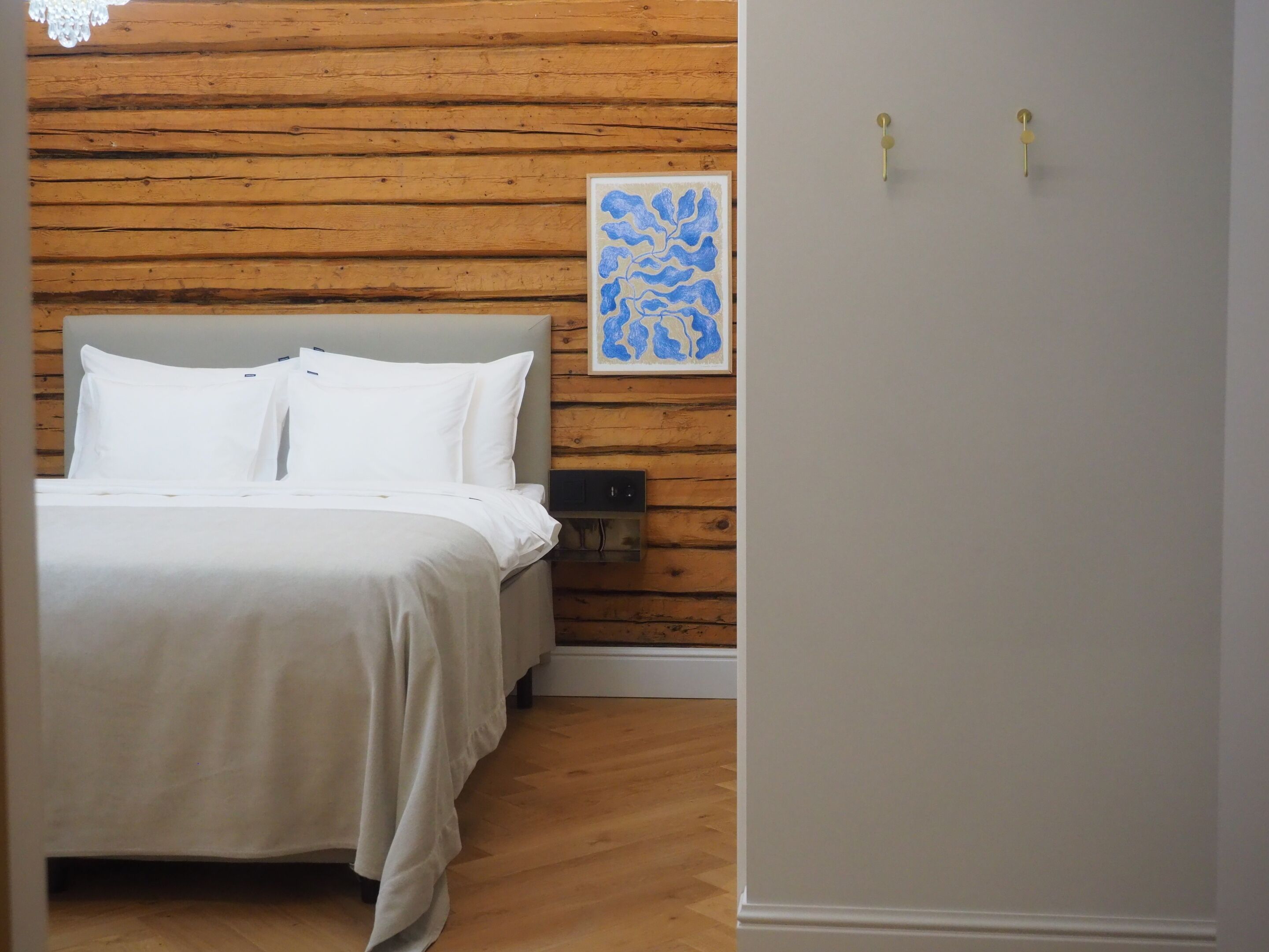 Un dormitorio con cabecera de madera, cama con ropa de cama blanca, mesita de noche, interruptor de luz en la pared y una obra de arte enmarcada.