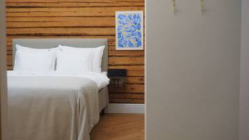 Un dormitorio con cabecera de madera, cama con ropa de cama blanca, mesita de noche, interruptor de luz en la pared y una obra de arte enmarcada.
