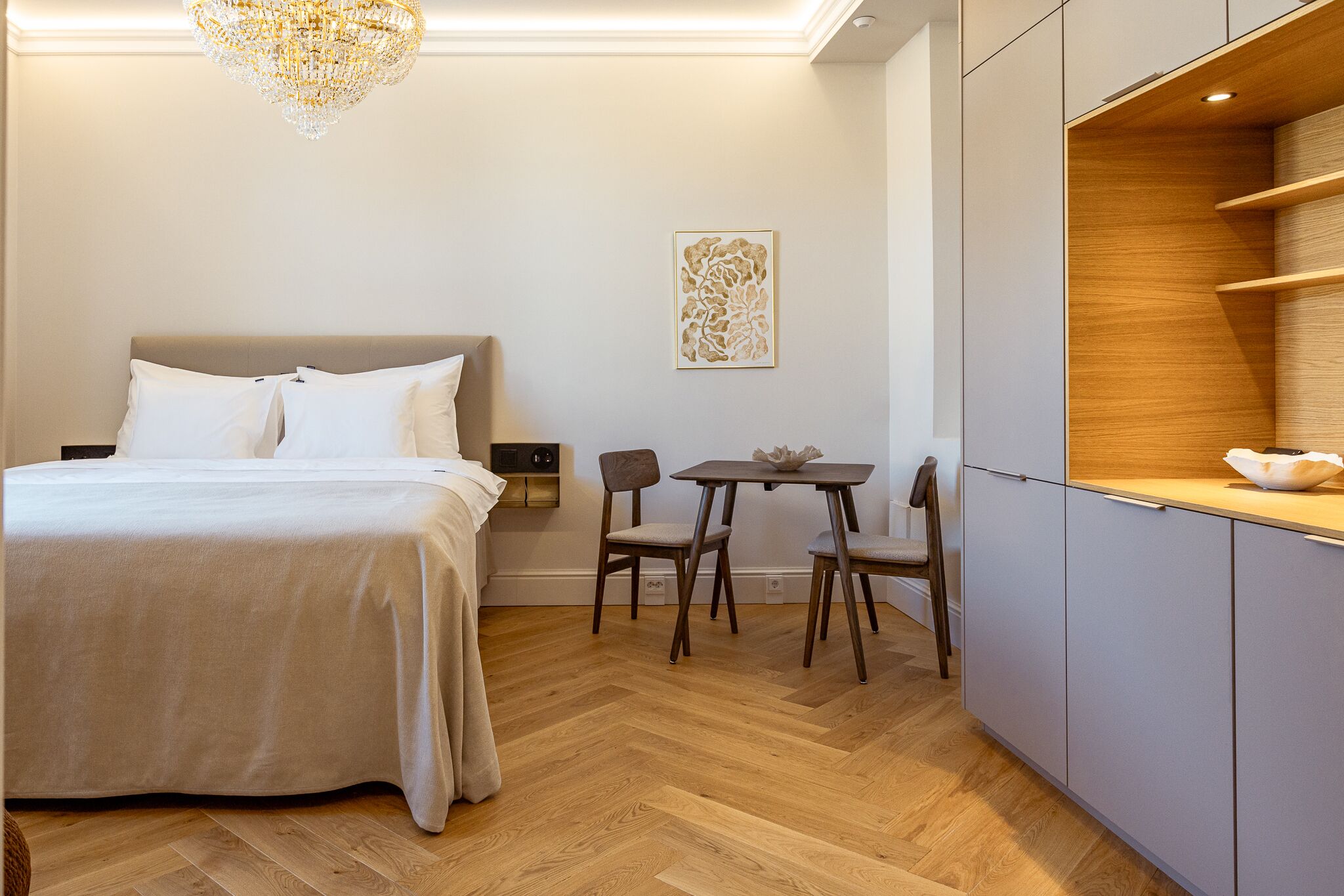 Una habitación de hotel con una cama, una pequeña mesa con sillas, un adorno en la pared y un armario de madera.
