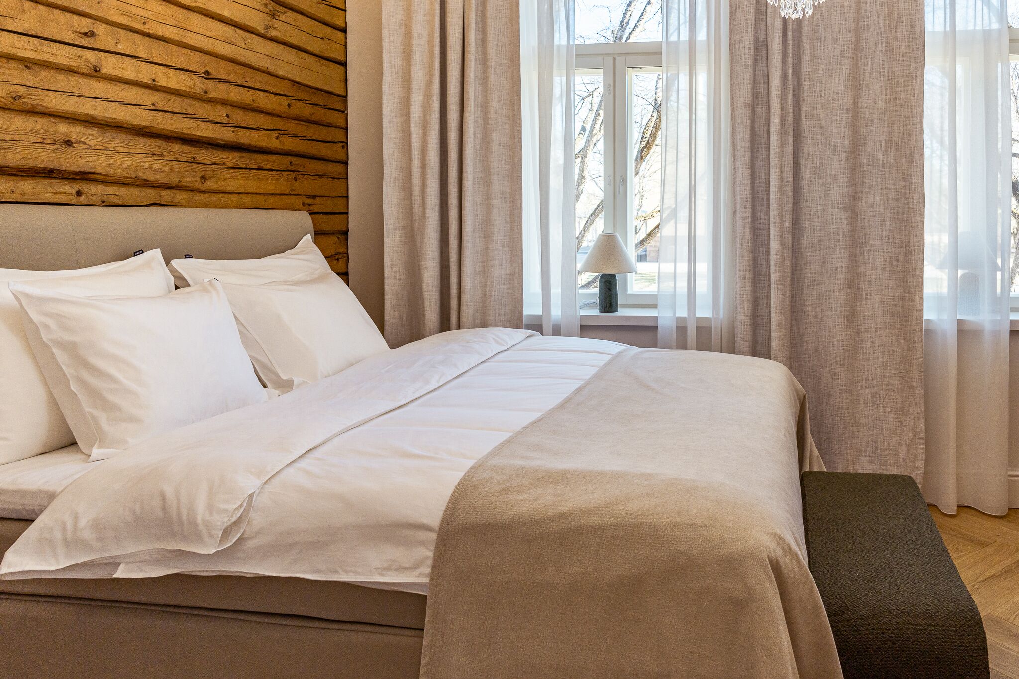 A bed with white bedding, a wooden headboard, and a view of the outdoors through sheer curtains.