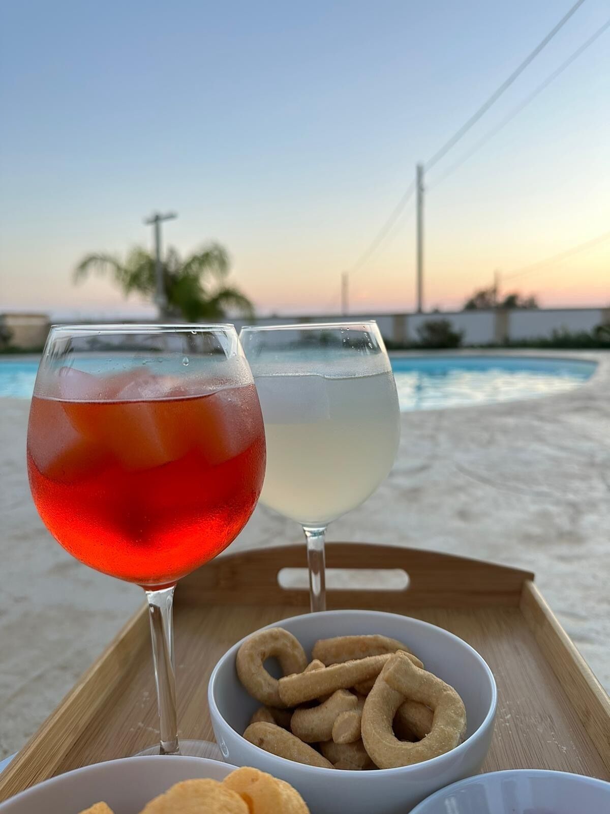 Dos vasos de bebidas sobre una bandeja con un bol de aperitivos y una piscina al fondo.