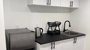 A compact kitchen with white cabinets, a stainless steel refrigerator, a black countertop, a kettle, a coffee maker, and a sink. - Room-haven in the Prairies 1BR B (Winnipeg)