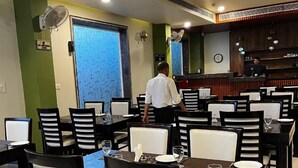 A restaurant with tables and chairs, a bar area, and a person in the background. - Sanskar Grand by Maan (Jaipur)