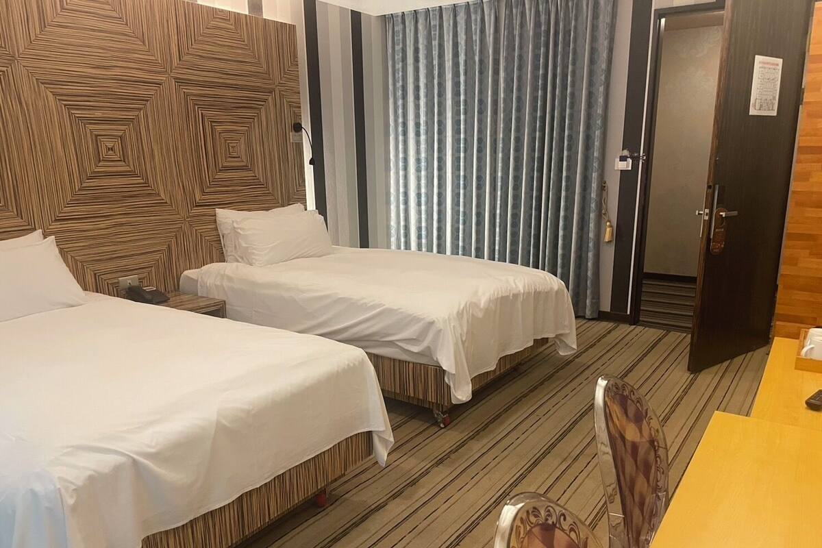 A hotel room with two beds, a wooden headboard, a striped carpet, a door, and a table with a chair.