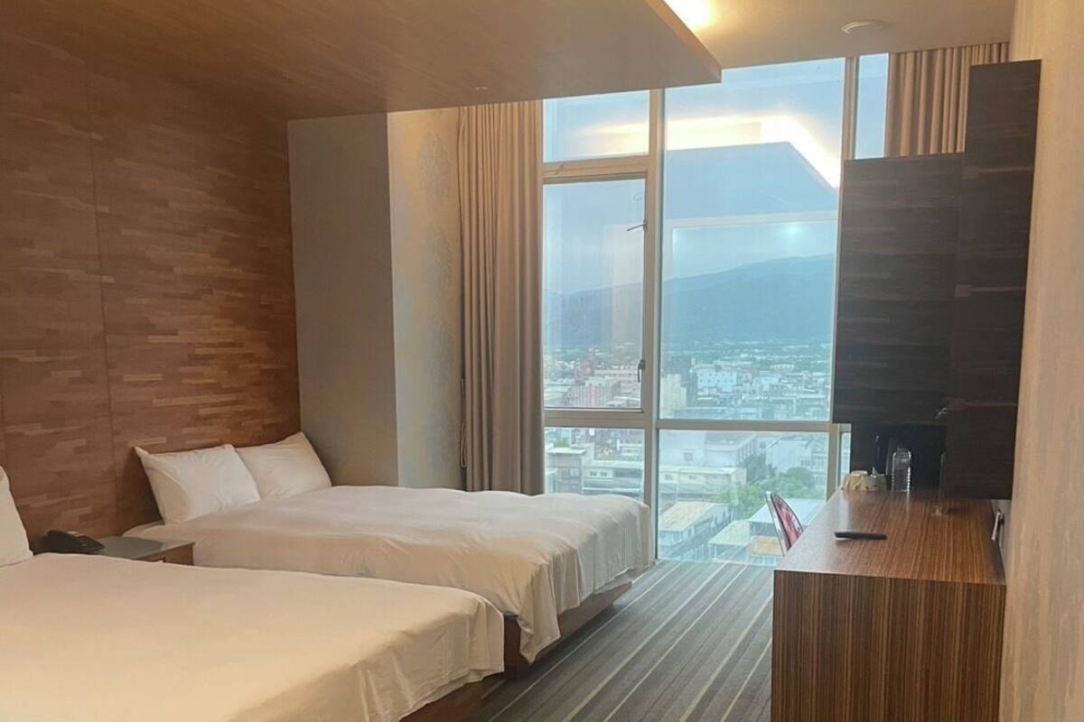 A hotel room with two beds, a desk, a trash bin, and a large window offering a city view.