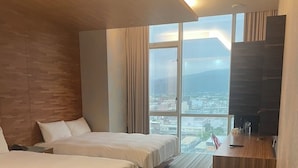 Ett hotellrum med två sängar, ett skrivbord, en papperskorg och ett stort fönster med utsikt över staden.