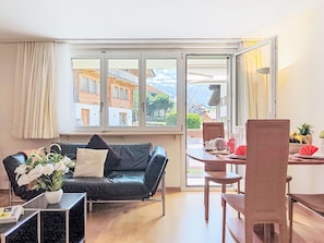 Un salon avec un canapé en cuir noir, une table à manger avec des chaises, une fenêtre donnant sur des immeubles et une plante en pot.