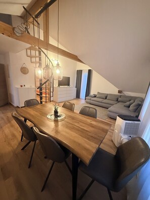 A dining area with a wooden table and chairs, a staircase, a living room with a sofa, and a TV. - GalleryLoft Wintertime Lake Thun&Mountainview (Thun)