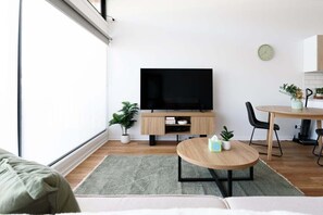 Una sala moderna con una televisión de pantalla plana, una mesa de centro de madera, un sofá y un comedor con una mesa pequeña y sillas.