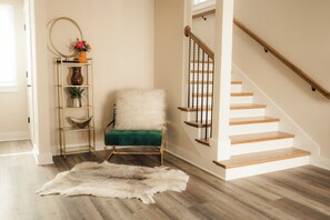 Una escalera con escalones y barandilla de madera, una butaca tapizada de verde, una alfombra de piel blanca y un estante metálico con objetos decorativos.