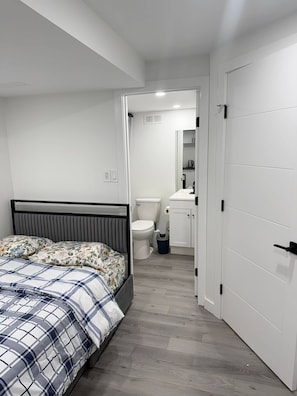 Un dormitorio con cama, un baño con inodoro y lavamanos, y una puerta blanca.