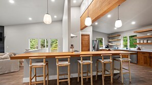 A modern kitchen with a long island, bar stools, wooden shelves, and a view of greenery. - Alaska Adventure Retreat LUXURY 4 Bed 5 bath in Girdwood, Base of Alyeska Resort (Anchorage)