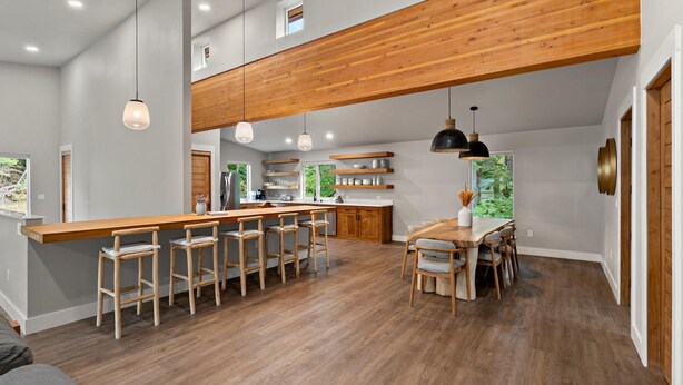 A spacious kitchen with a wooden bar, bar stools, a dining table with chairs, pendant lighting, and a view of greenery outside. - Alaska Adventure Retreat LUXURY 4 Bed 5 bath in Girdwood, Base of Alyeska Resort (Anchorage)