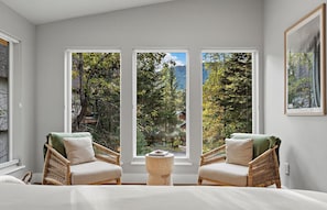 A bedroom with a large window, two wicker chairs, a small wooden stool, and a bed.