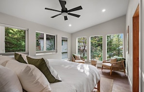 A bedroom with a large bed, a ceiling fan, and multiple windows offering a view of trees.