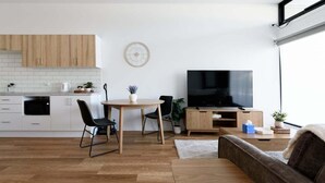 A modern kitchen with wooden cabinets, a dining area with a table and chairs, a sofa, and a flat-screen TV. - Minimalist Studio Near Burnie | Clean & Cozy (Hillcrest)