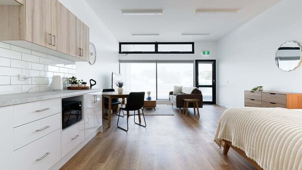 A modern kitchen with white cabinets, a dining area with a table and chairs, a sofa, and a bed with a white cover. - Minimalist Studio Near Burnie | Clean & Cozy (Hillcrest)