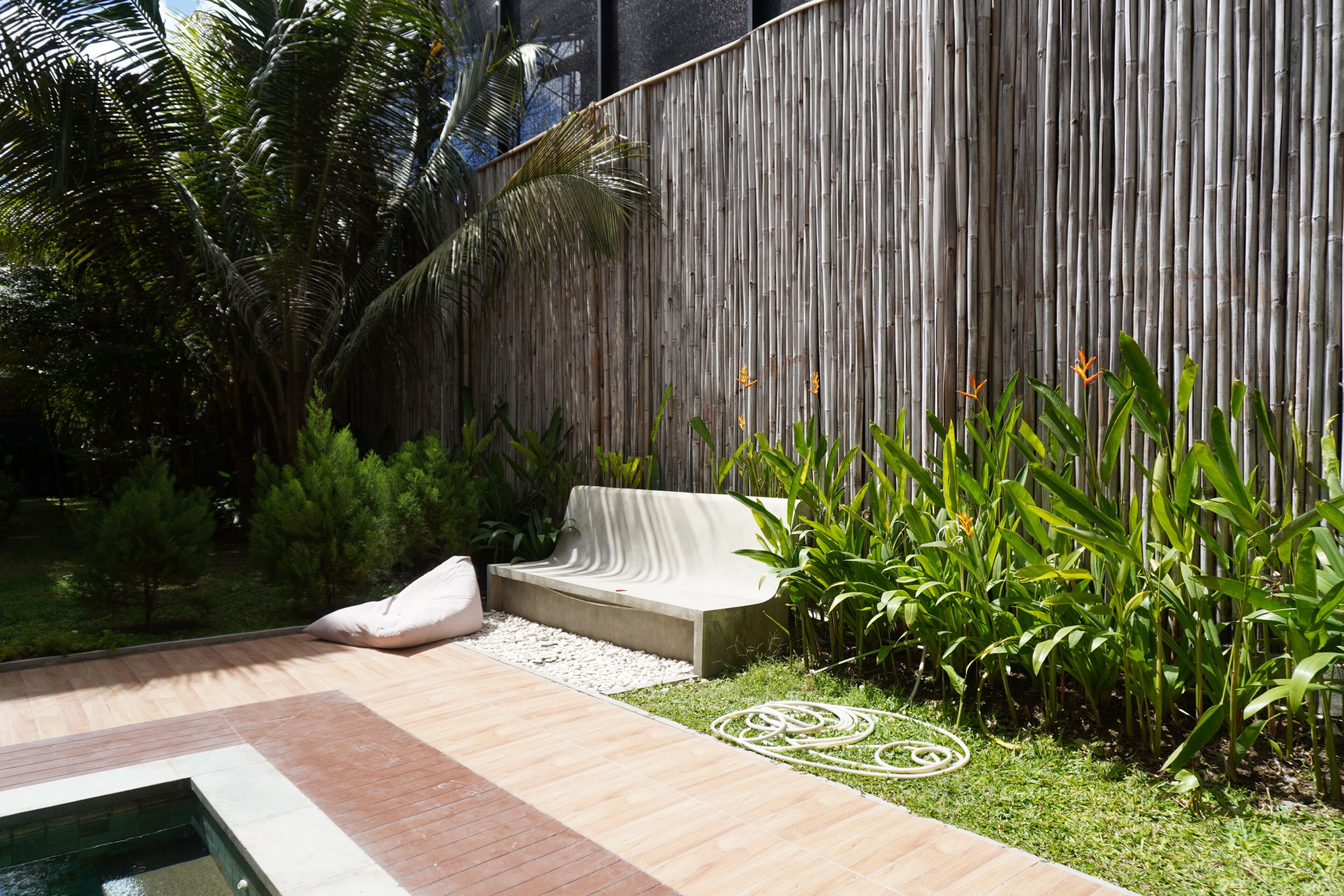 A modern outdoor seating area with a concrete bench and a white cushion, surrounded by lush greenery and a bamboo fence.