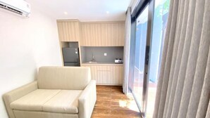 A modern living room with a sofa, a kitchen area with cabinets and a refrigerator, and a large sliding glass door. - Pamela Bali (Keroboken Kelod)