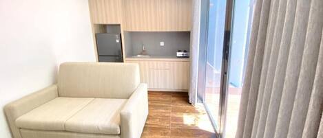 Una sala de estar moderna con un sofá, un área de cocina con armarios y un refrigerador, y un gran ventanal corredero.
