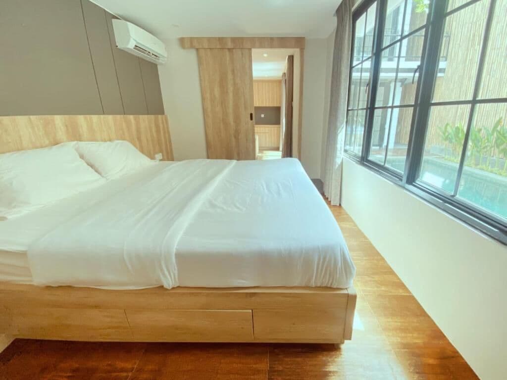 A bed with a wooden headboard, a window with a view, and an air conditioning unit.