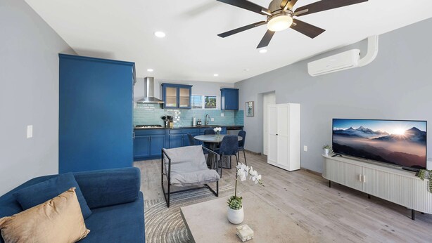 A modern living room with a sofa, TV, ceiling fan, and a kitchen in the background. - Broadway Casita - Remodeled Minutes from U of A (Tucson)
