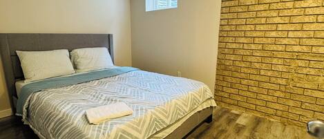 Un dormitorio con cama, ventana, pared de ladrillo y puerta.