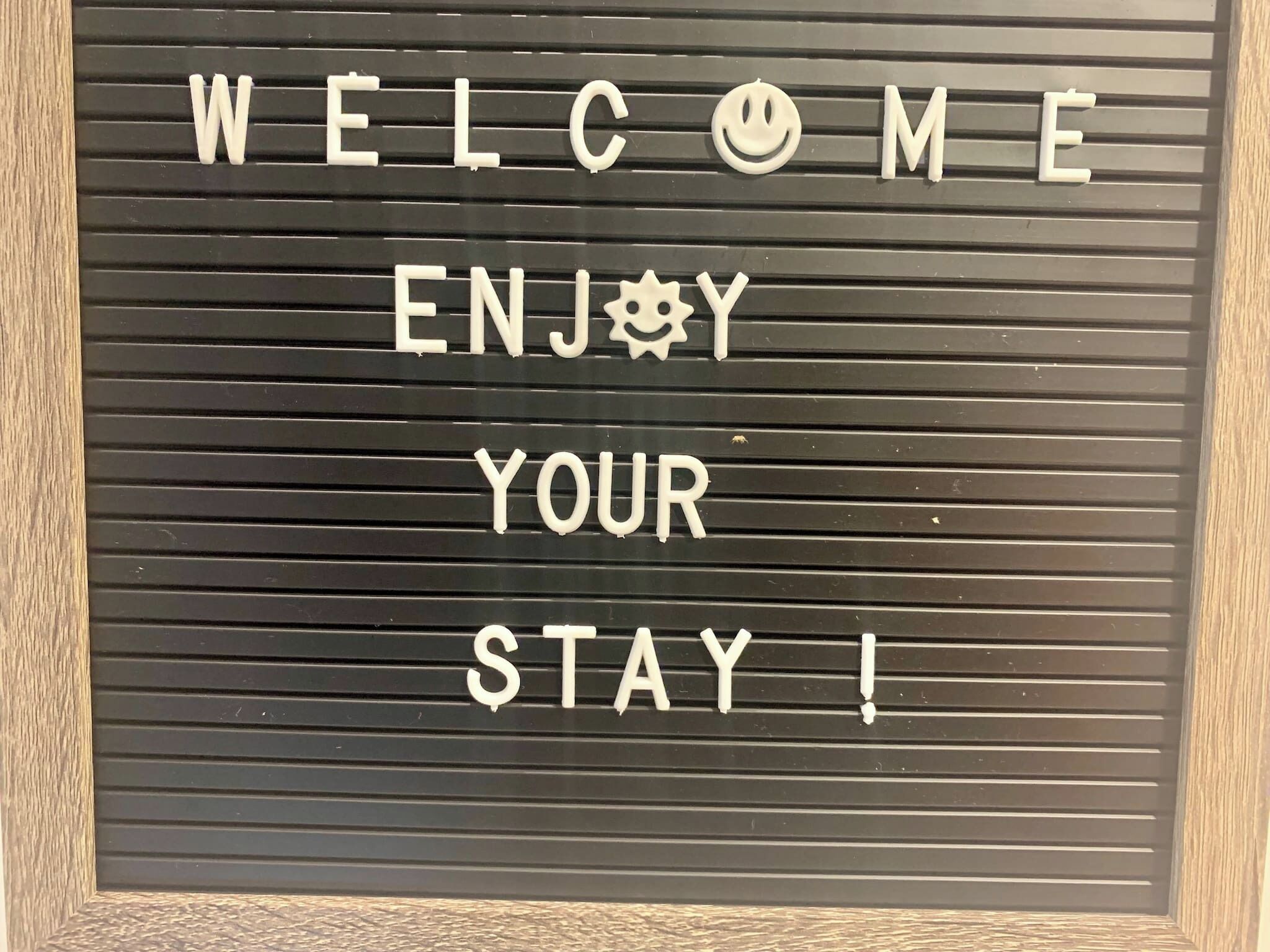 Placa de boas-vindas com o texto 'WELCOME ENJOY YOUR STAY!' em fundo preto com linhas horizontais.