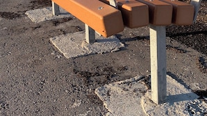 A bench with a metal frame and wooden slats, set on a gravel surface with a clear sky and distant buildings in the background. - Room-haven in the Prairies Full 2BR (Winnipeg)