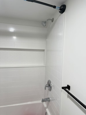 Ducha de azulejos blancos con cabezal de ducha de mano, barra de ducha manual y un perchero negro.