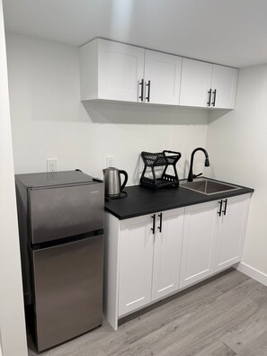 A compact kitchen with white cabinets, a stainless steel refrigerator, a kettle, a coffee maker, and a sink with a faucet. - Room-haven in the Prairies Full 2BR (Winnipeg)