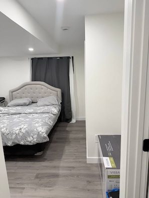 A bedroom with a bed, grey curtains, a white wall, and a wooden floor. - Room-haven in the Prairies Full 2BR (Winnipeg)