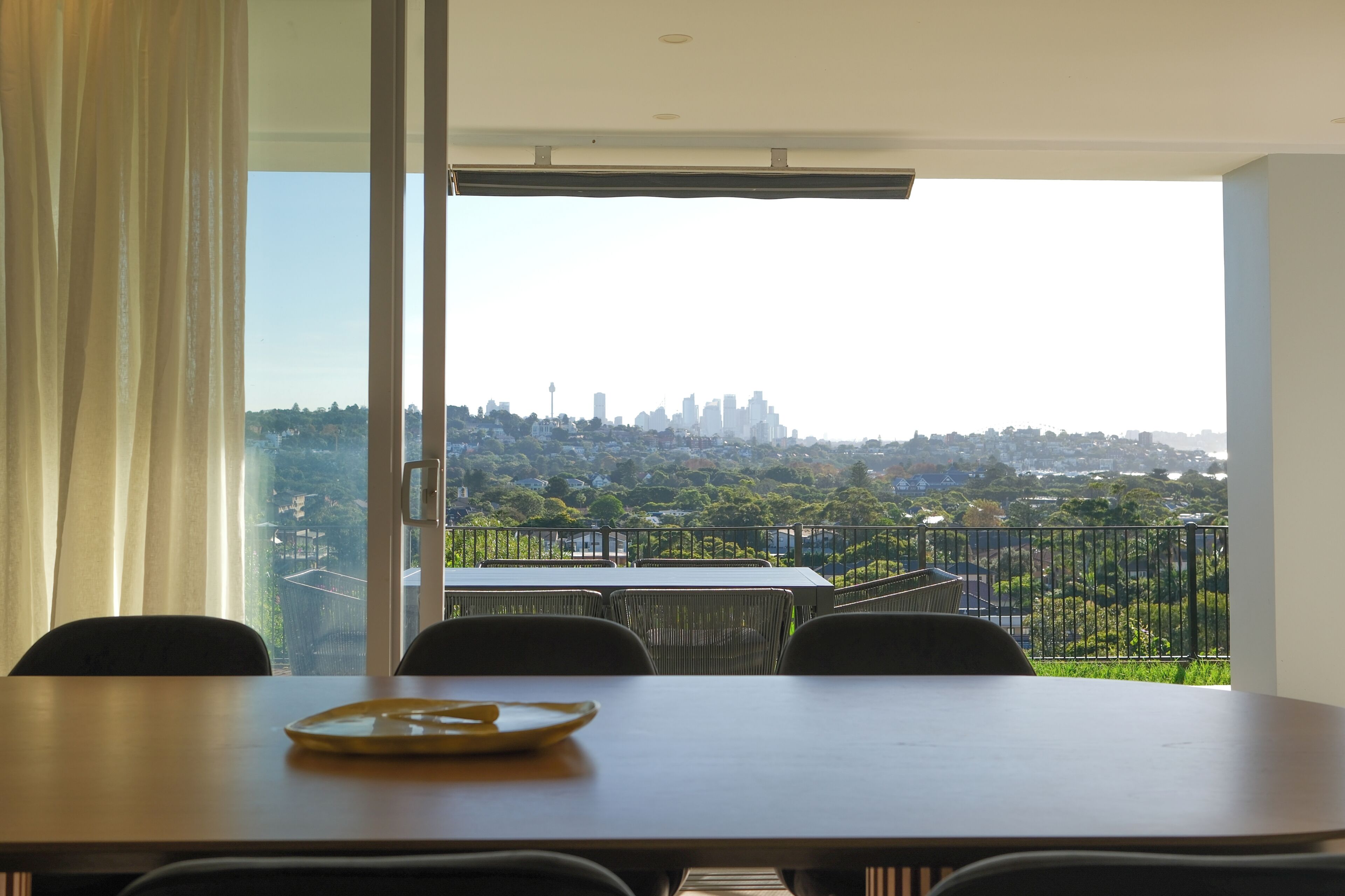 A dining area with a table and chairs, a view of a cityscape through a balcony, and a large window with curtains.