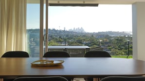 Un comedor con una mesa y sillas, vistas de la ciudad a través de un balcón y un amplio ventanal con cortinas.
